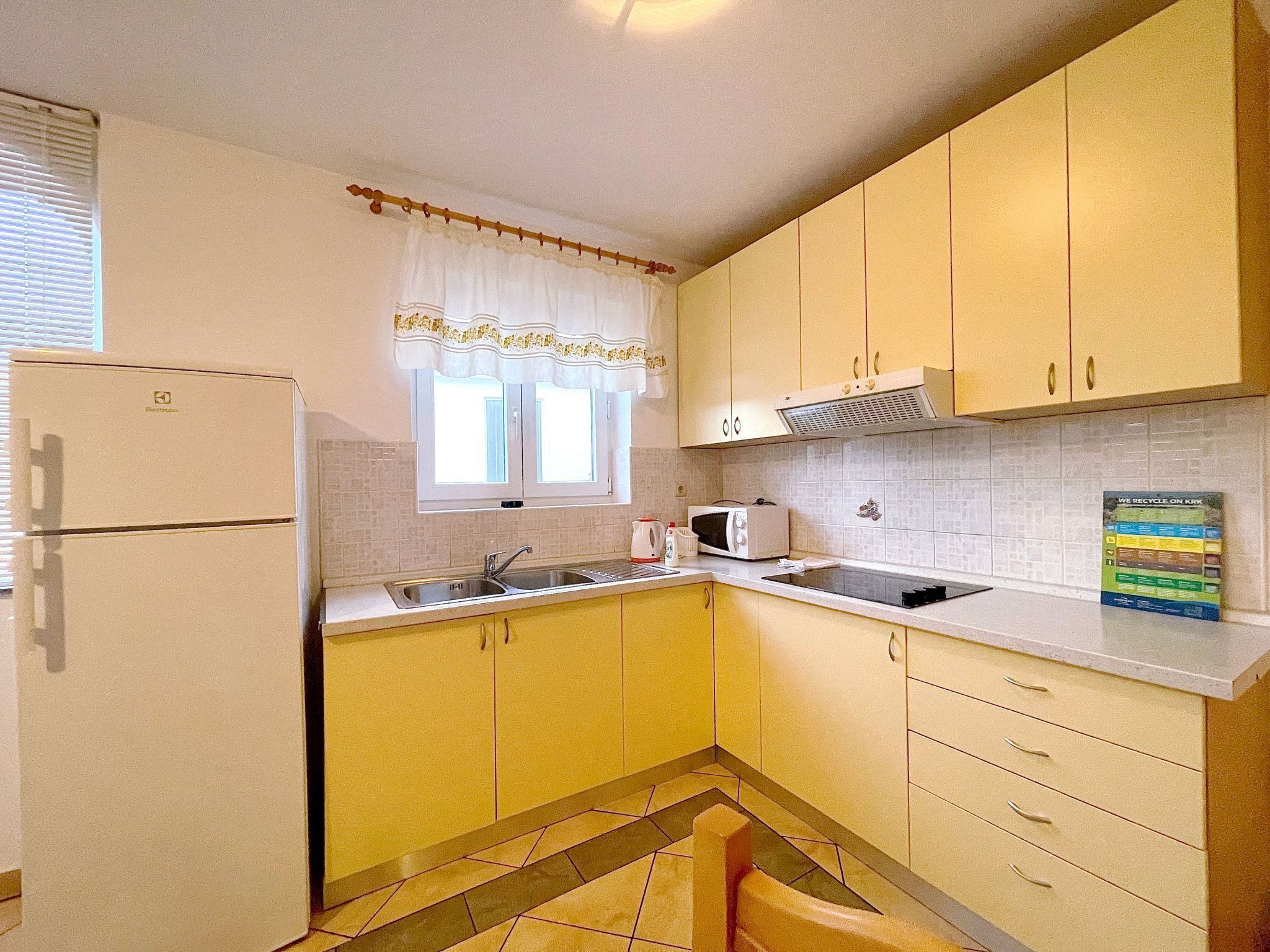A compact kitchen with a refrigerator, sink, microwave, and a window with curtains.