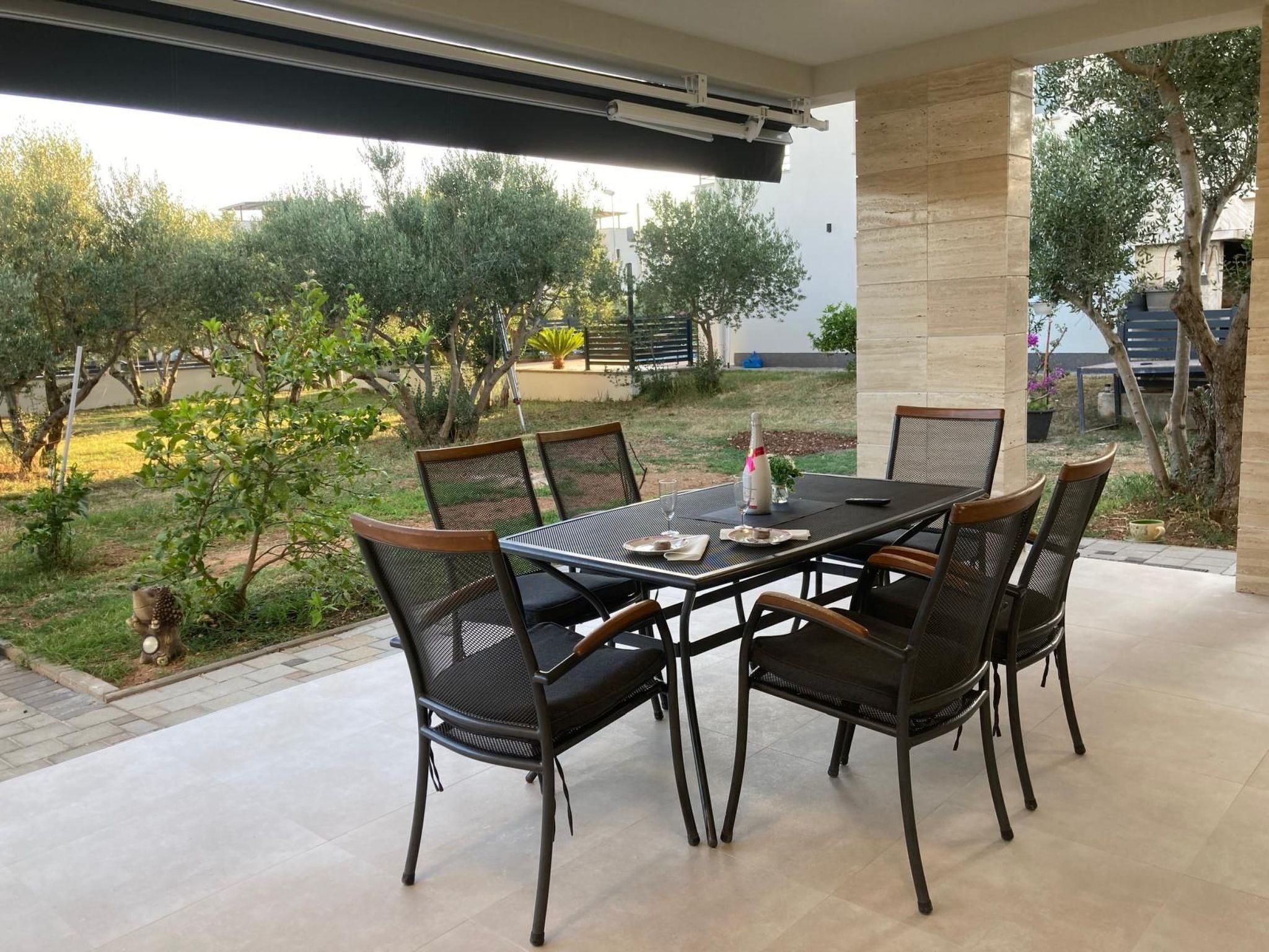 A covered patio with a table and chairs, surrounded by greenery and a view of the outdoors.
