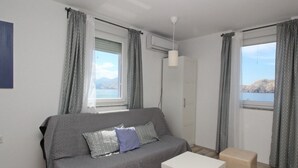 Ein modernes Wohnzimmer mit einem grauen Sofa, einem weißen Tisch und einem beigefarbenen Hocker. An den Fenstern hängen Vorhänge mit Muster, und an der Wand ist eine Klimaanlage zu sehen.