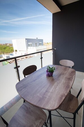 Ein Balkon mit Holztisch und -stühlen, einer violetten Pflanze und Blick auf Gebäude im Hintergrund.