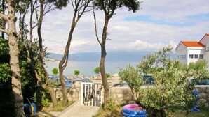 Un sentier de pierres mène à un mur de pierres surmonté d’une porte, le tout étant entouré d’oliviers et offrant une vue sur la mer.