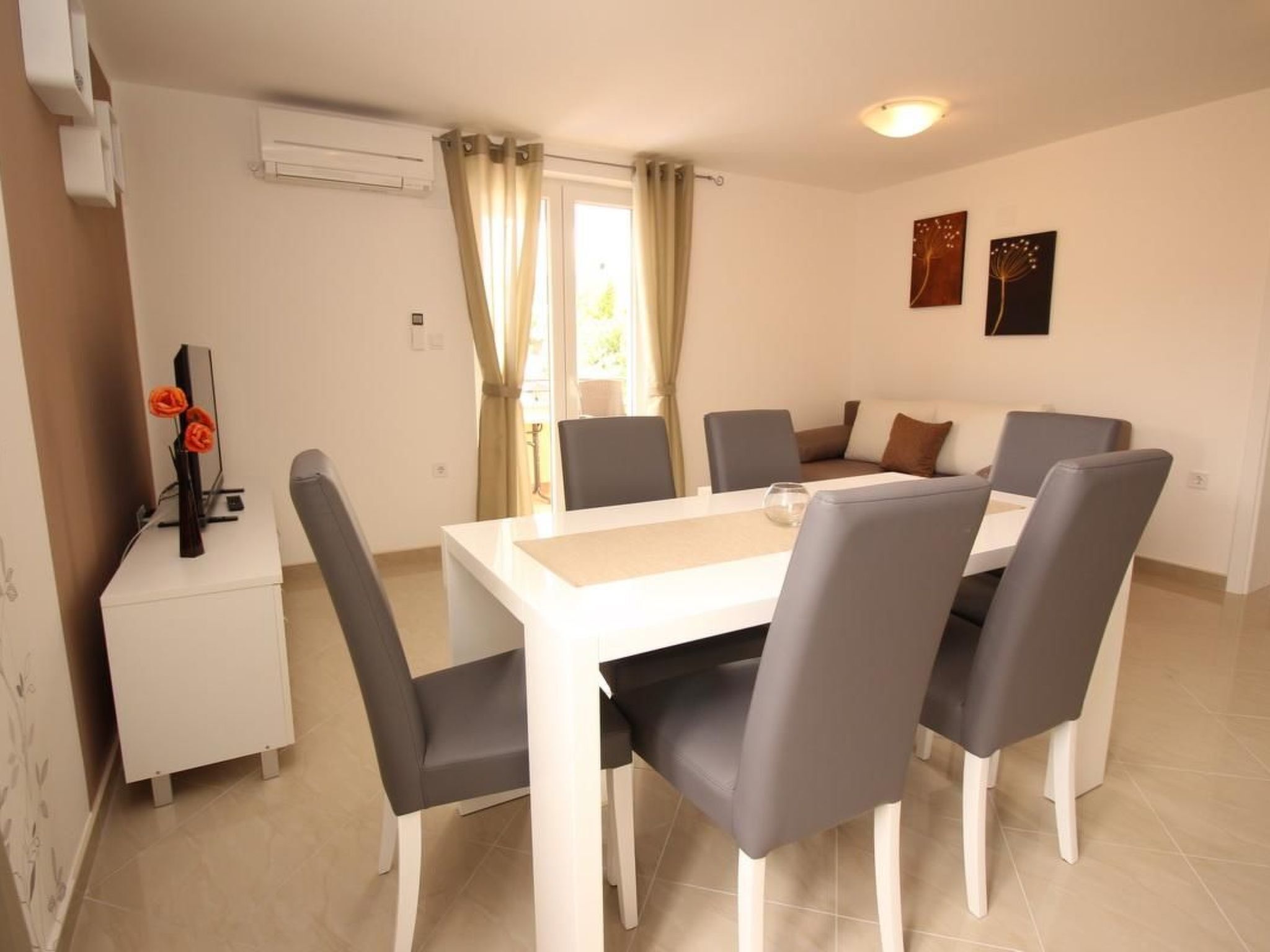 Un comedor con una mesa blanca y sillas grises, un pequeño armario blanco con un florero, y una ventana con cortinas.