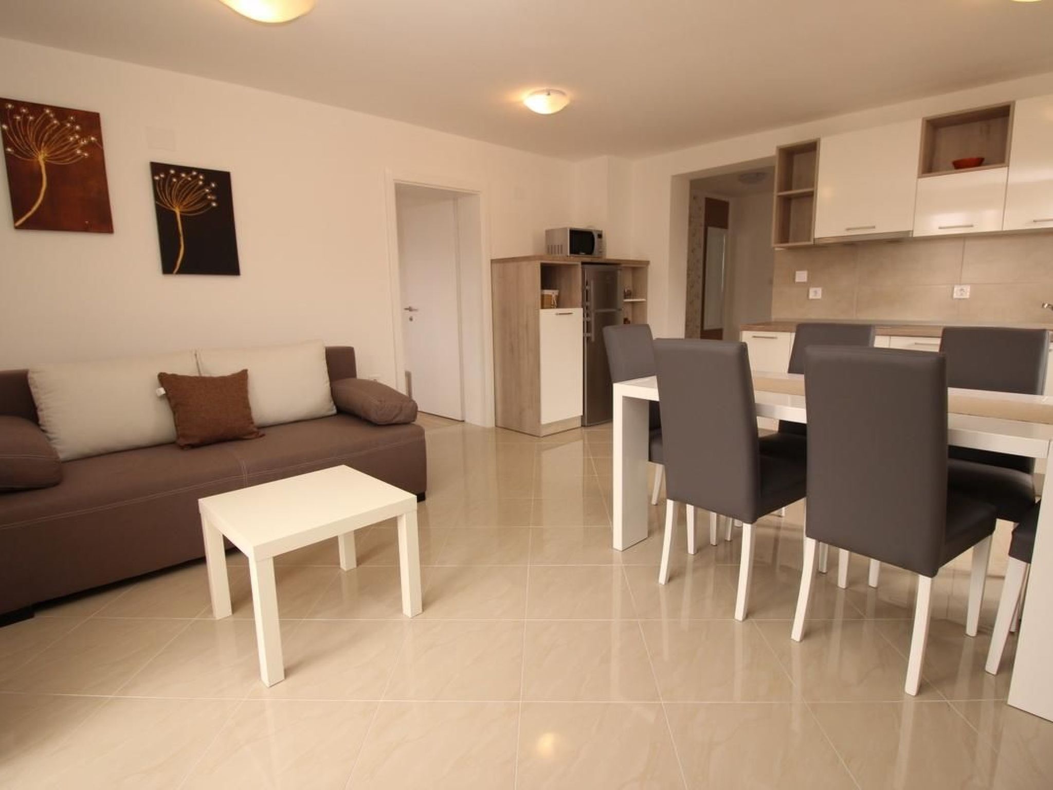 A modern living room with a brown sofa, white coffee table, and black chairs. There is a dining area with a white table and chairs. The room has white walls and a tiled floor.