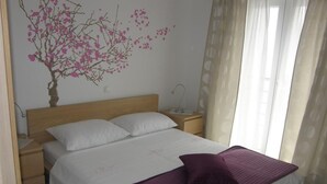 Ein ordentlich bezogenes Bett mit weißer und violetter Bettwäsche, ein hölzerner Kopfteil, ein Nachttisch mit Lampe und eine Wanddekoration mit rosa Blumen.