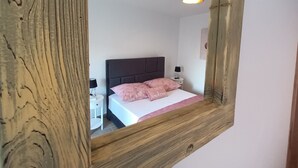 Une chambre à coucher avec un miroir à cadre en bois, un lit avec une literie blanche et des oreillers roses, une table de chevet avec une lampe et une petite commode.