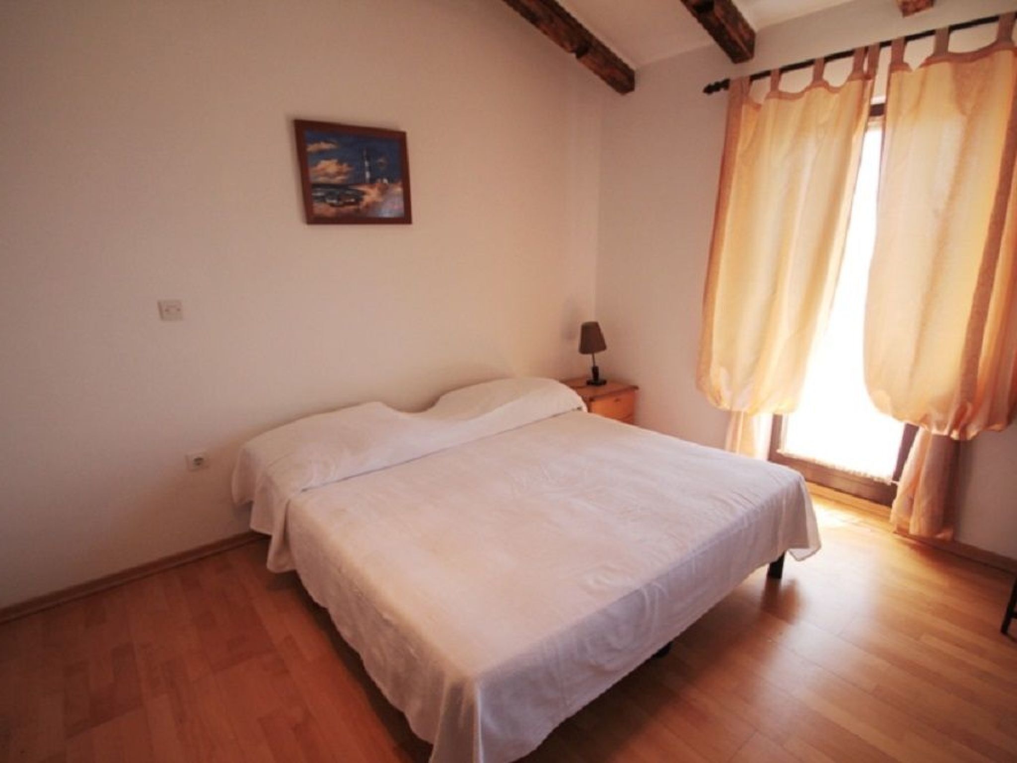 A bedroom with a bed, a nightstand, a lamp, a painting on the wall, and a window with curtains.