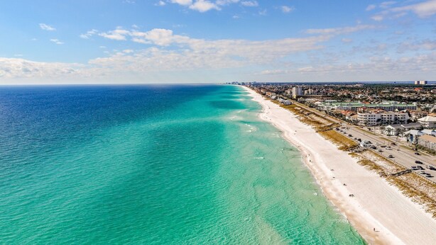 Un paisaje urbano costero con una extensa playa de arena, aguas turquesas cristalinas y una zona urbana densa con edificios y carreteras.