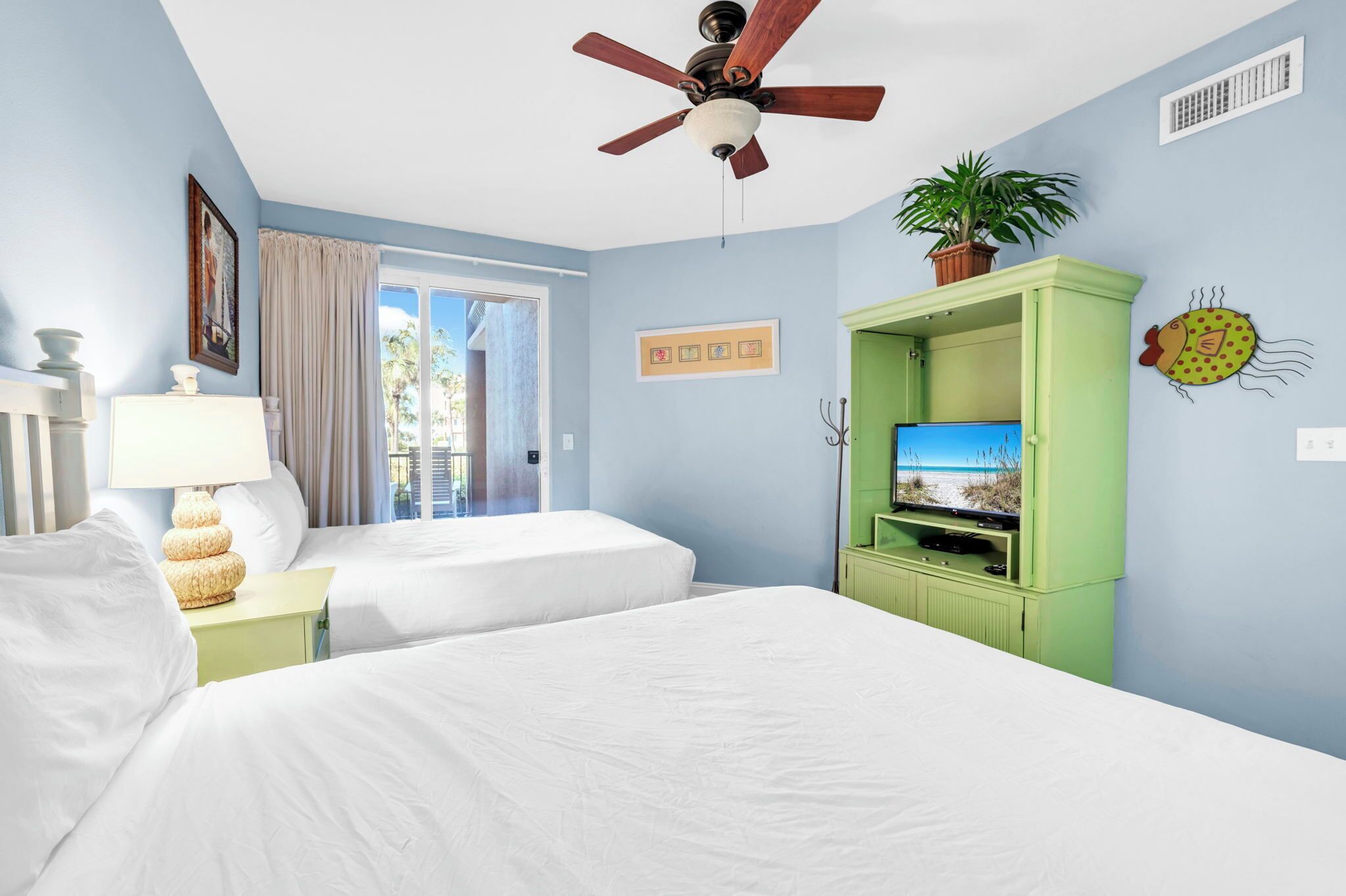 Habitació d'hotel amb dos llits, un ventilador de sostre, un televisor sobre un armari verd i una finestra amb cortines.