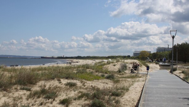 Une plage de sable avec une promenade en bois, des dunes herbeuses et une étendue d’eau.