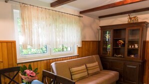 Una habitación acogedora con sofá, una mesa de comedor con mantel floreado, un aparador con puertas de cristal y una ventana con cortinas traslúcidas.