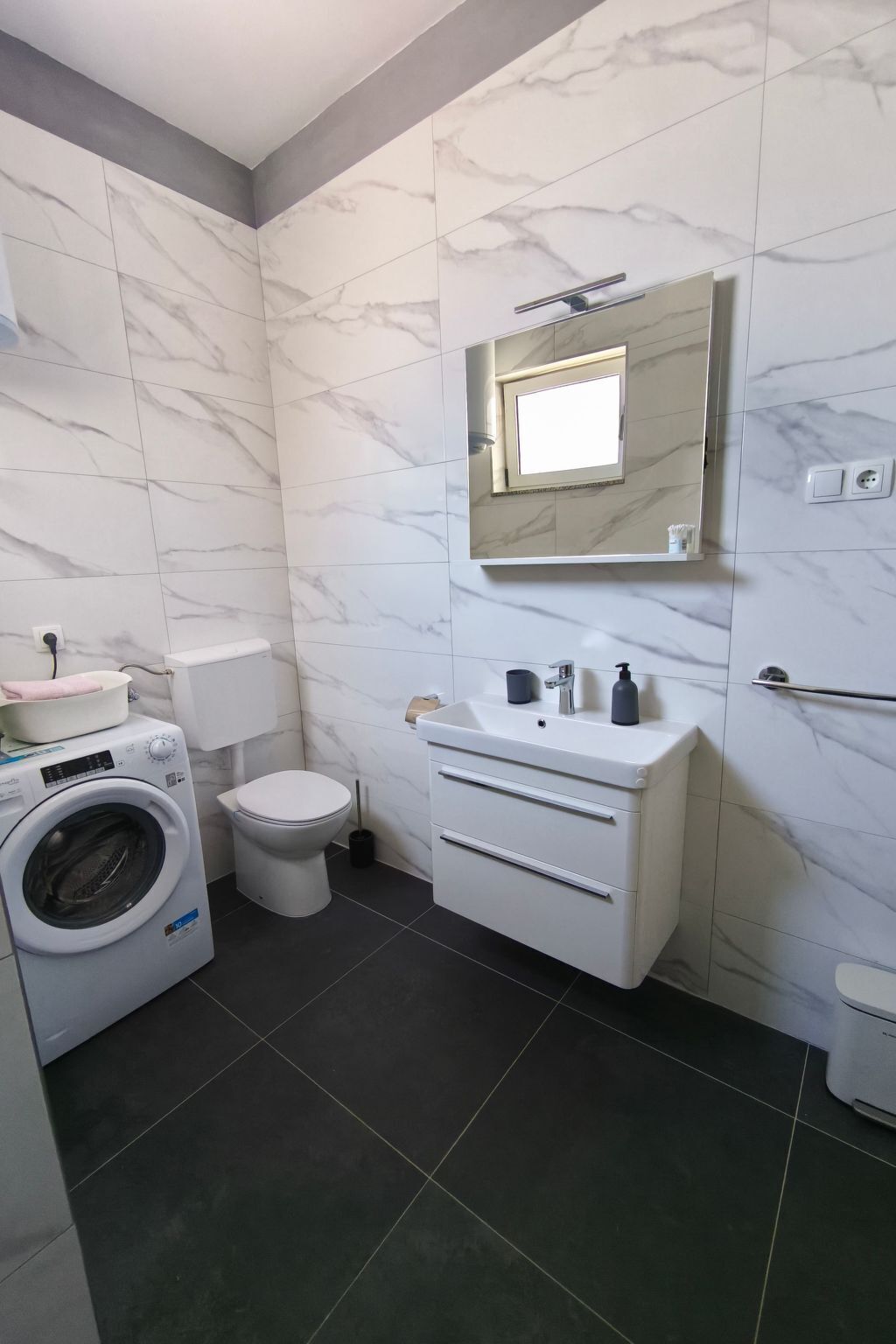 Un baño moderno con pared de mármol, lavadora integrada, inodoro y tocador con espejo.