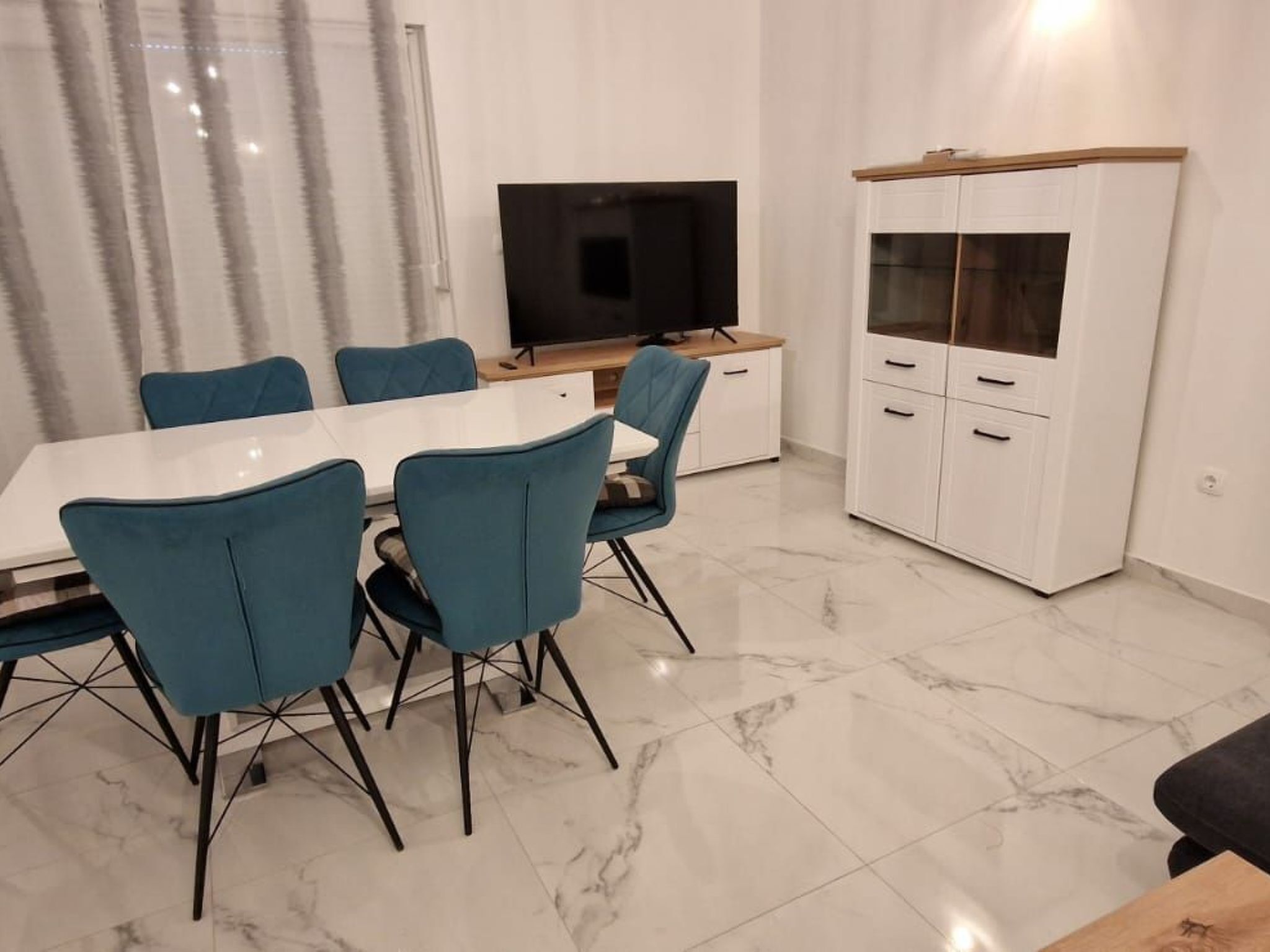 A modern dining area with a white table and teal chairs, a TV on a wooden stand, and a white cabinet with glass doors.
