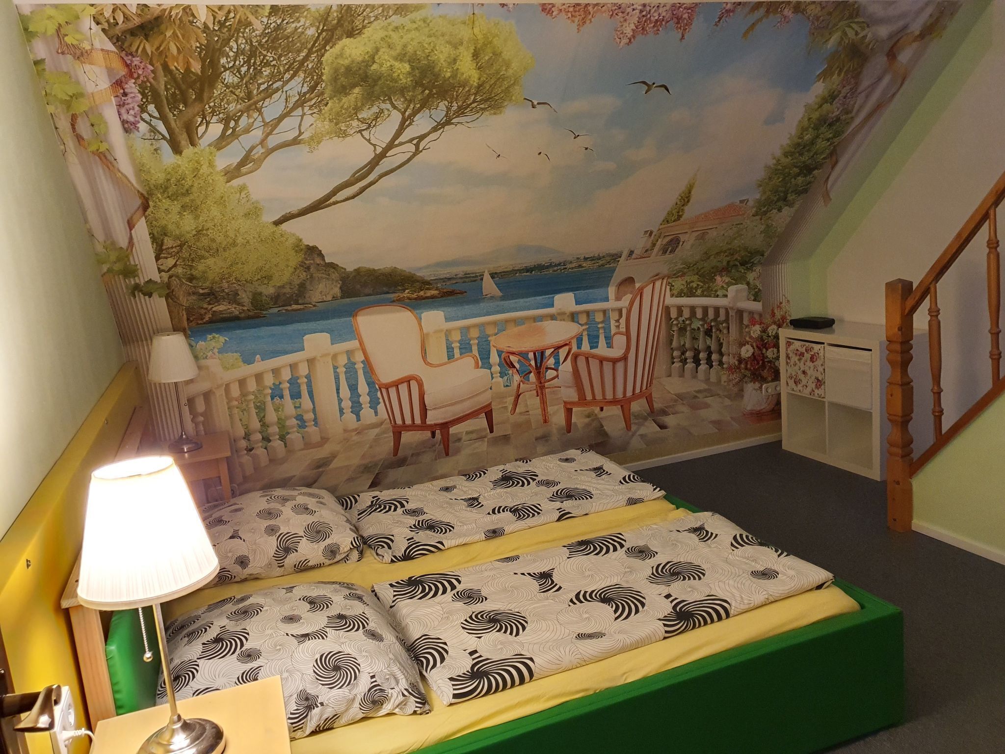 A bedroom with a mural of a seaside scene, a bed with patterned bedding, a nightstand, and a lamp.