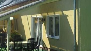 Ein gelbes Gebäude mit einem geneigten Dach, einer überdachten Veranda mit Stühlen und einem kleinen Garten.