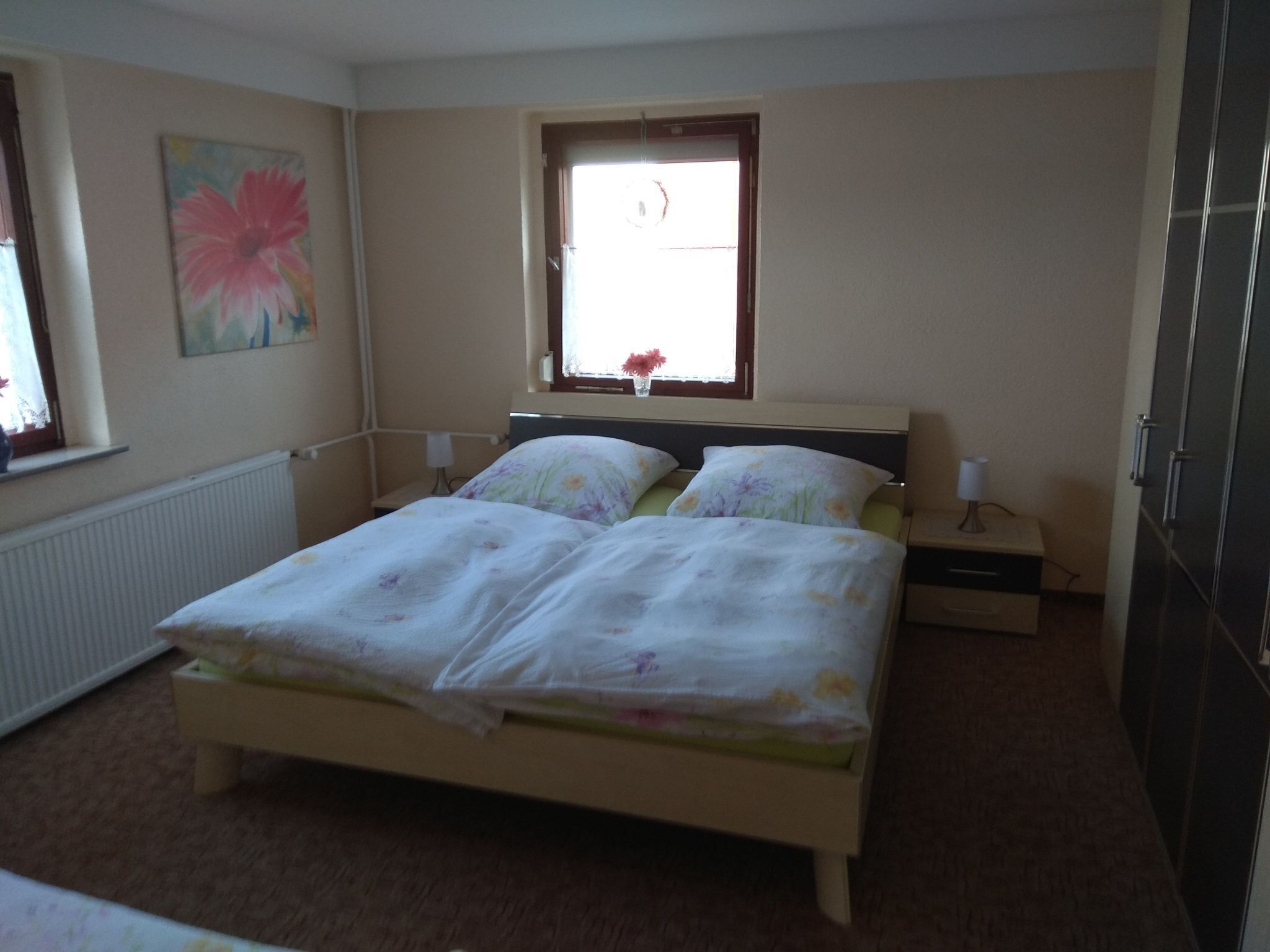 Une chambre à coucher avec un lit, une table de chevet, une lampe, un radiateur, une fenêtre avec une plante et un tableau au mur.