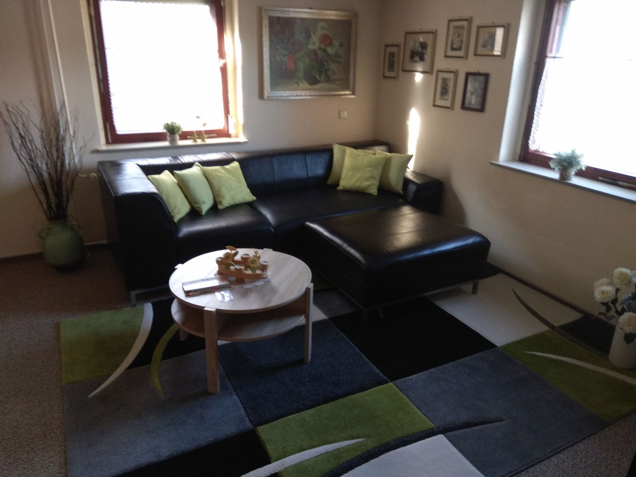 En stue med en sort sofa, grønne puder, et rundt sofabord og en gulvtæppe med et mønster.