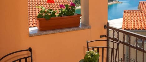 A balcony with a table and chairs, a window with a view of the sea and mountains, and a potted plant on the table.