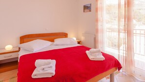 A neatly made bed with white linens and a red blanket, a bedside lamp, and a view of a balcony through sheer curtains. - Wohnung "Apartman für 4 Personen" by Interhome (Brela)