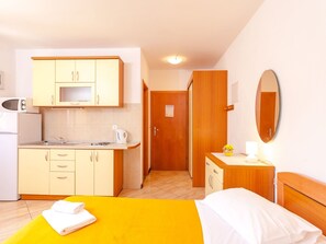 Une chambre d’hôtel compacte comprenant un coin cuisine, un lit avec une couvre-lit jaune, une table de chevet avec une lampe et un miroir au mur.