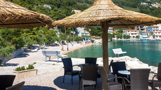 A beachside café with wicker chairs and tables, a thatched umbrella, and a view of the sea and mountains in the background. - Studio im Erdgeschoss für zwei Personen mit einer Terrasse by Interhome (Brela)