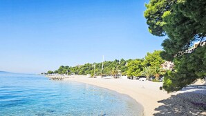 A coastal beach with clear blue waters, pebbly shore, and a backdrop of trees and buildings. - Studio im Erdgeschoss für zwei Personen mit einer Terrasse by Interhome (Brela)
