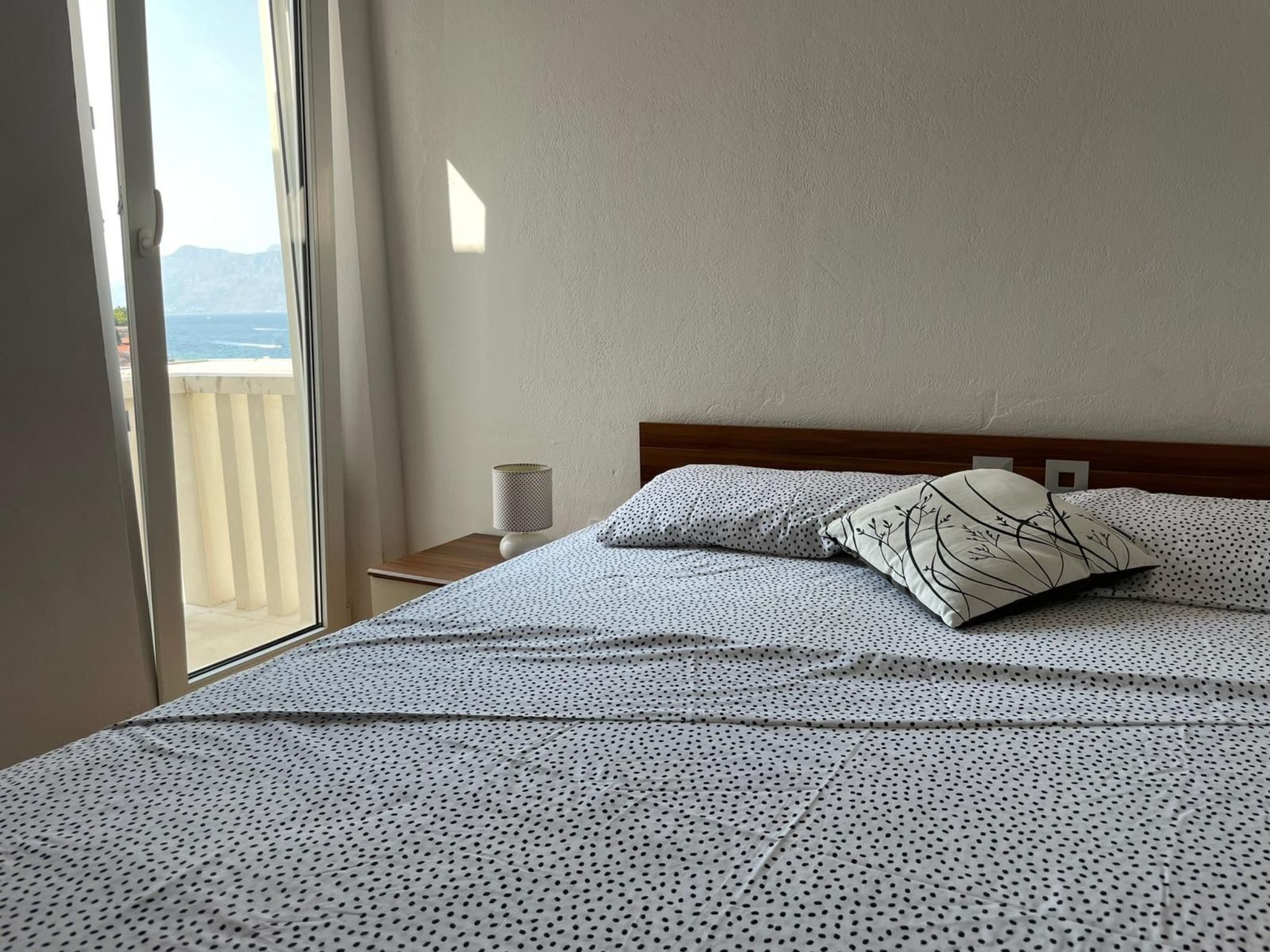A bedroom with a bed, a nightstand, a lamp, and a view of the sea through a window.