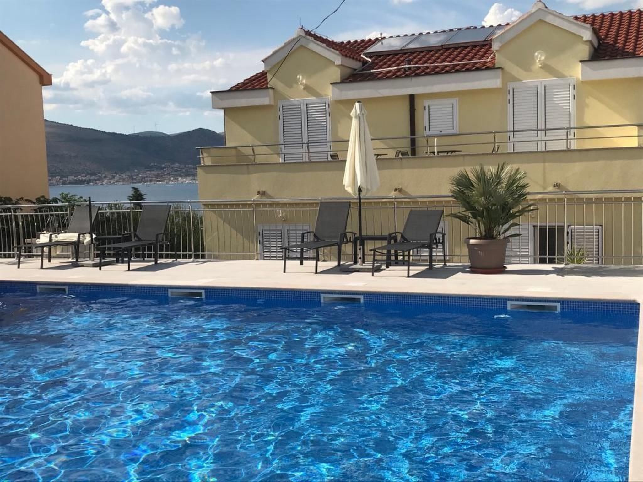 A swimming pool with lounge chairs and a potted plant, surrounded by a building with a tiled roof and a view of the sea.