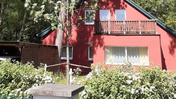 A red two-story house with a balcony, surrounded by trees and greenery.