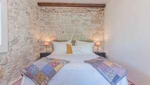Une chambre avec un mur en pierre, un plafond en bois, un lit avec une literie blanche, deux tables de chevet avec des lampes et une couverture colorée.