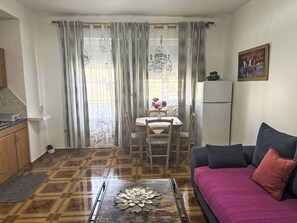 A living room with a sofa, a dining table with chairs, a kitchen area, and a window with decorative curtains. - Wohnung Antica bei Queen beach Nin by Interhome (Nin)