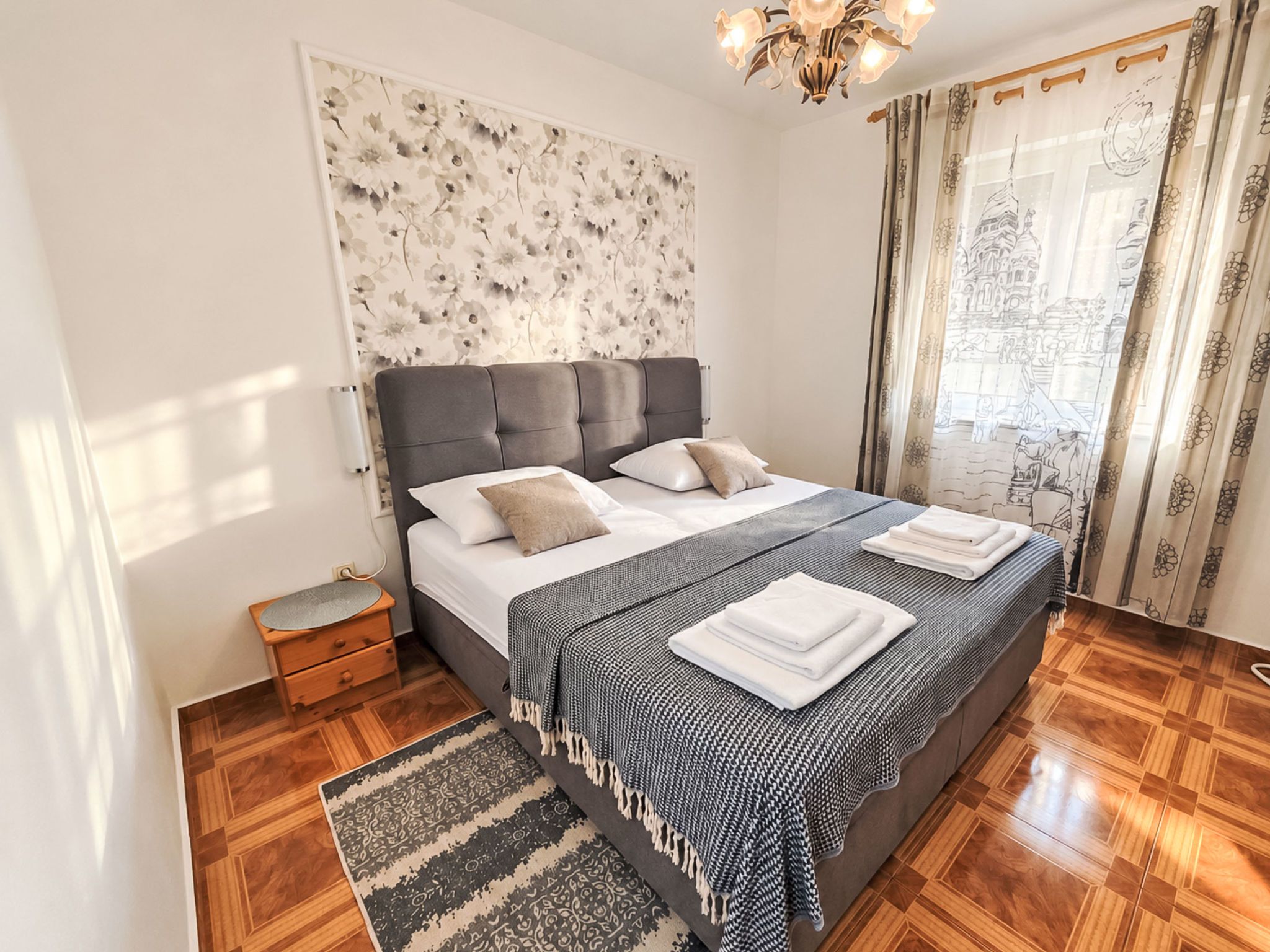 A bedroom with a large bed, a nightstand, a patterned rug, and a wall with a floral design.
