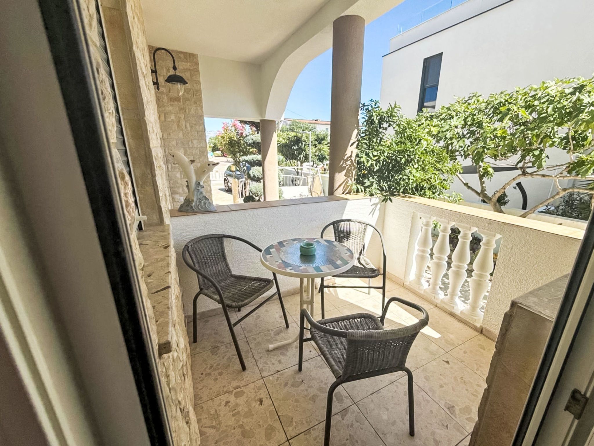 A balcony with two wicker chairs and a small round table, a stone pillar, and a view of a building and trees.
