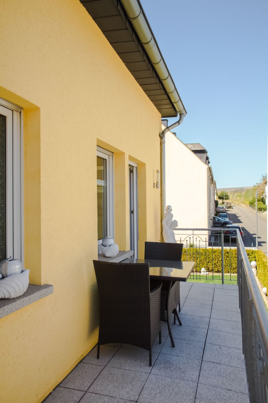 Ein Balkon mit Tisch und Stühlen, einer Statue und Blick auf eine Straße und Bäume.