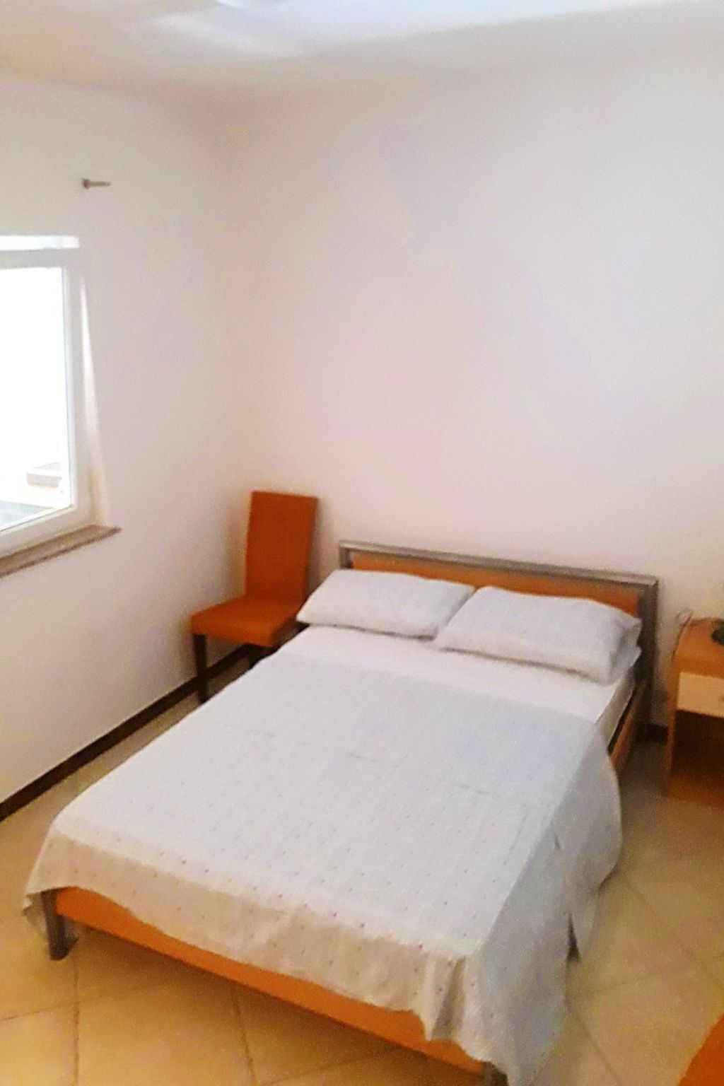 A bedroom with a bed, an orange chair, a nightstand, and a window.