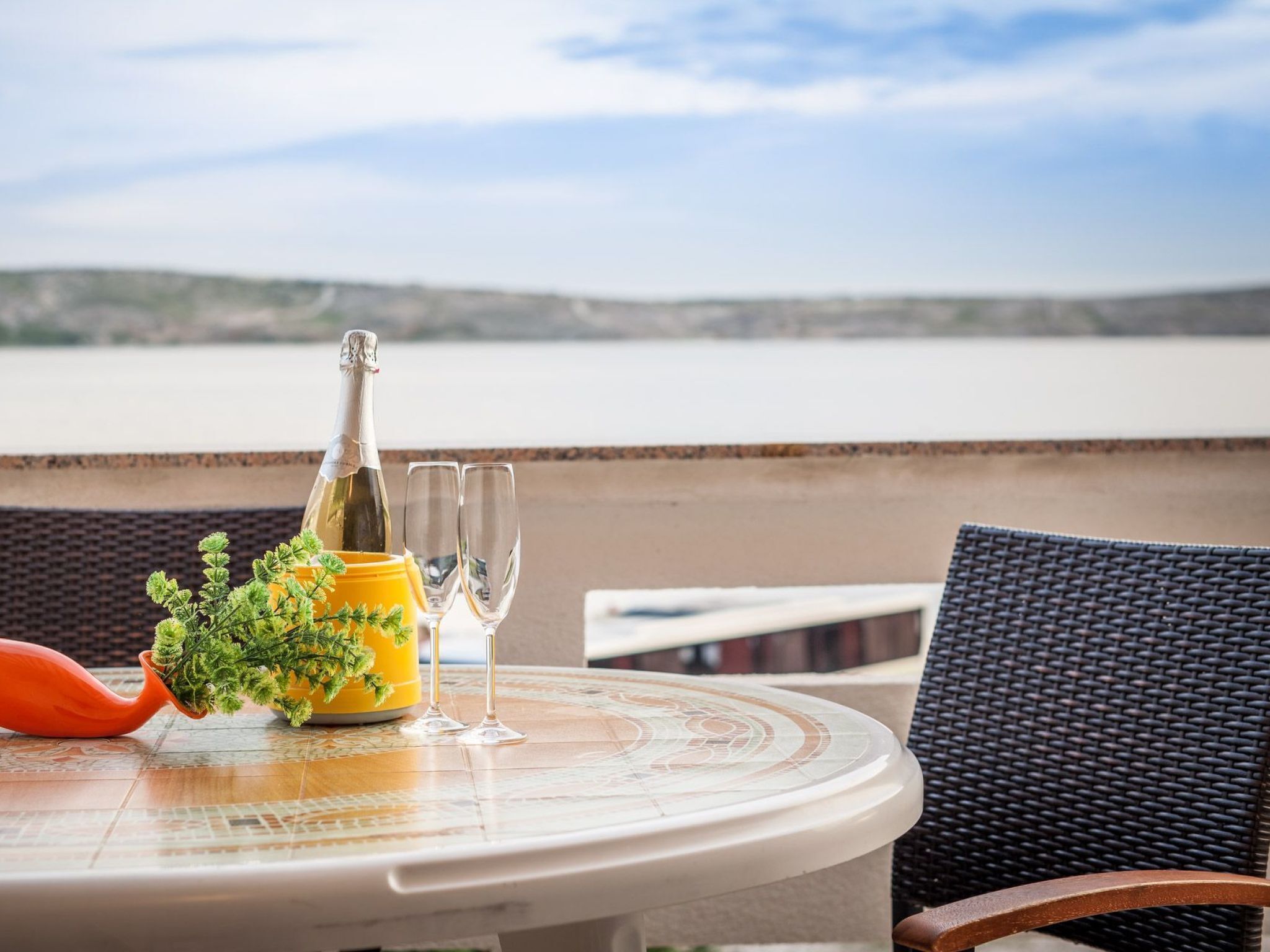 Een tafel met een champagnefles, twee glazen en een bloempot, op een balkon met uitzicht op een wateroppervlak.