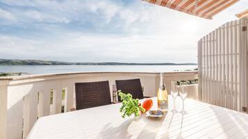 Une table dressée pour deux personnes, avec une bouteille de champagne, des verres et un panier de fruits, sur une terrasse donnant sur un plan d’eau.