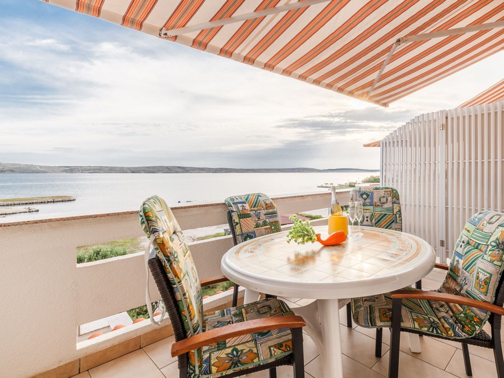 A round table with four chairs, a bottle, and a glass on a balcony overlooking a body of water.