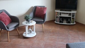 Un soggiorno con due poltrone grigie, un tavolino bianco con soprammobili, una TV a schermo piatto su un mobile bianco e una libreria.
