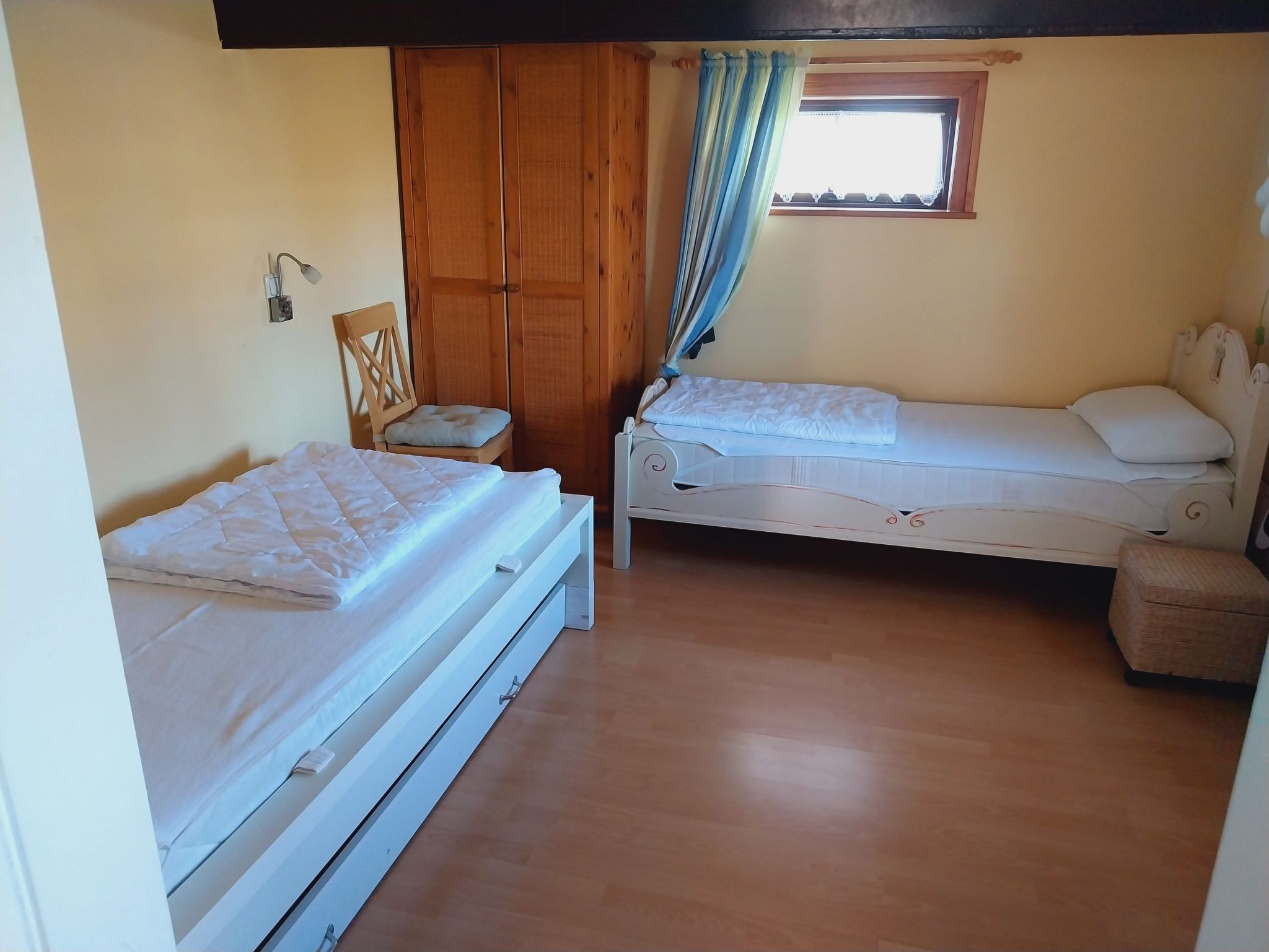 Una habitación con dos camas, un armario de madera, un ventanal con cortinas y una silla.