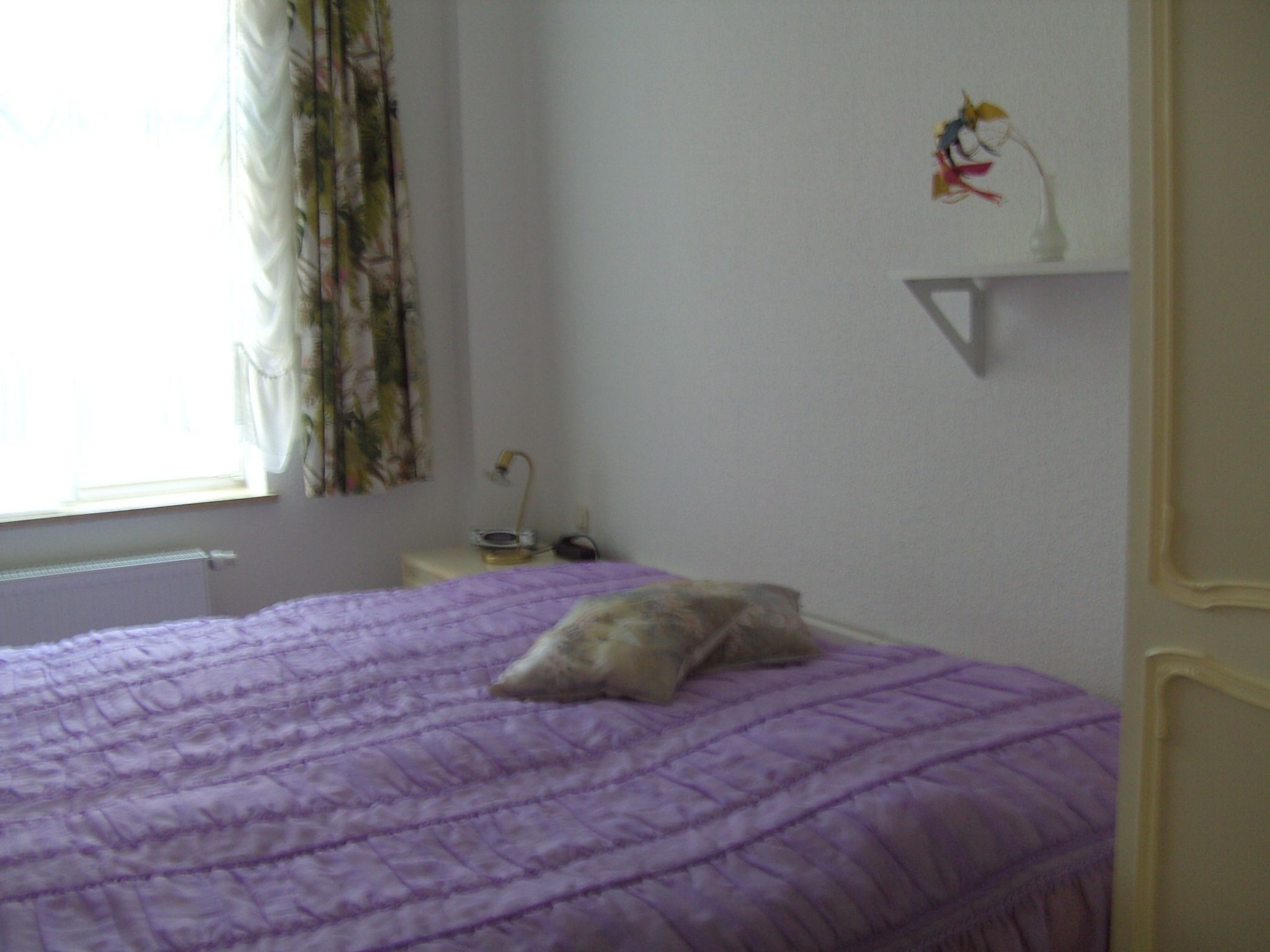 A bedroom with a purple bedspread, a pillow, a bedside table with a lamp, and a window with curtains.