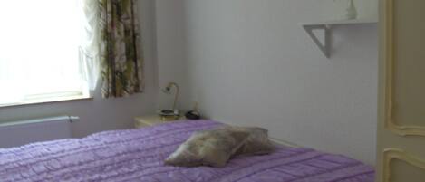 A bedroom with a purple bedspread, a pillow, a bedside table with a lamp, and a window with curtains.