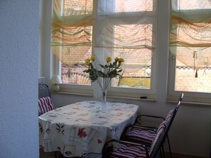 Et spiseområde med et bord dækket af et blomstret duge, en vase med gule blomster og udsigt til en murstensmur igennem vinduet.