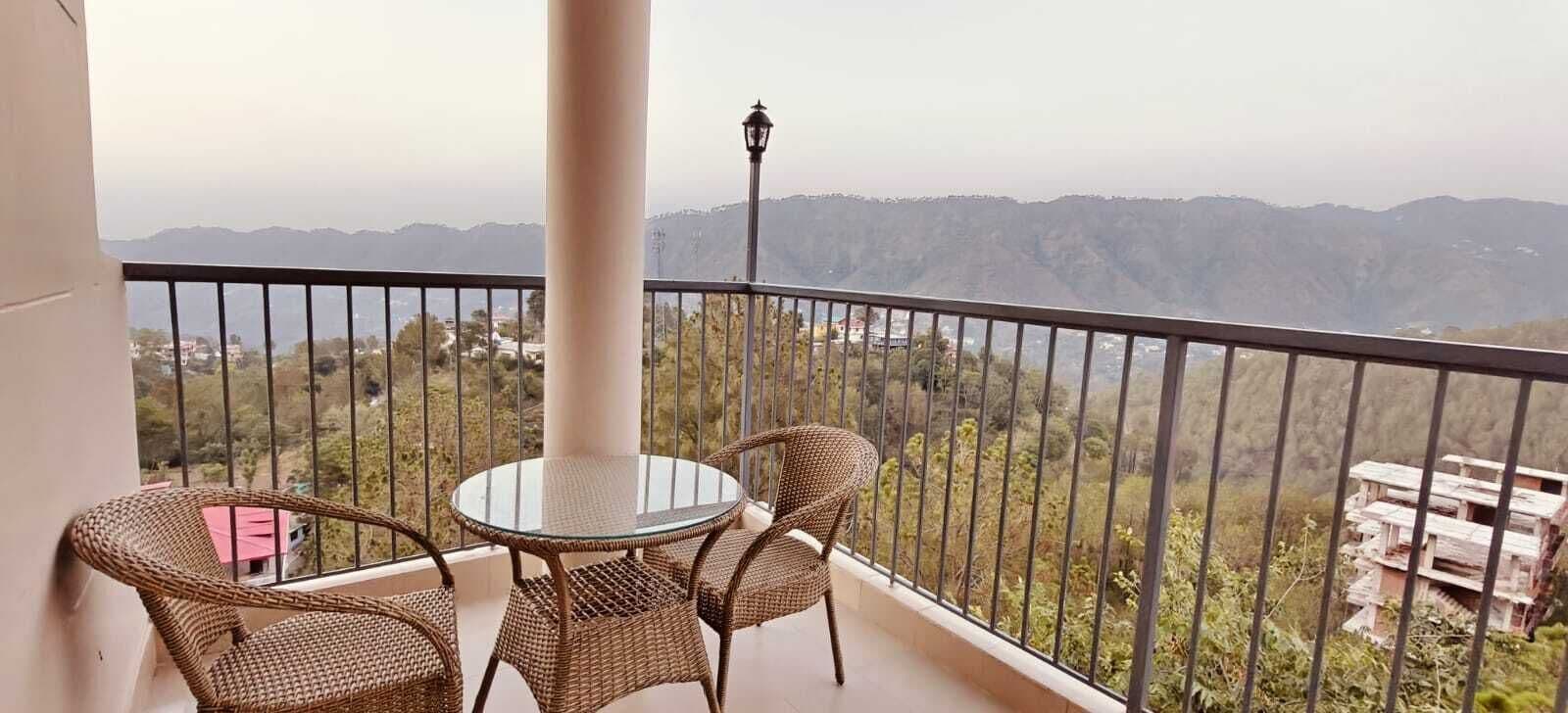 A balcony with a round glass table and wicker chairs, overlooking a mountainous landscape.