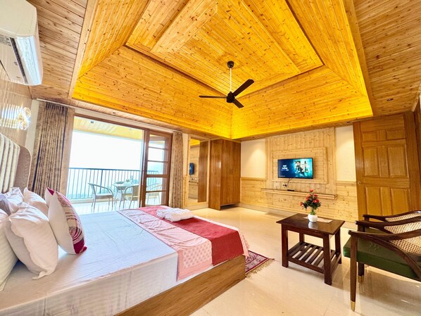 A hotel room with a bed, a TV, a ceiling fan, a small table, and a chair. - The Royal Terrace (Kasauli)