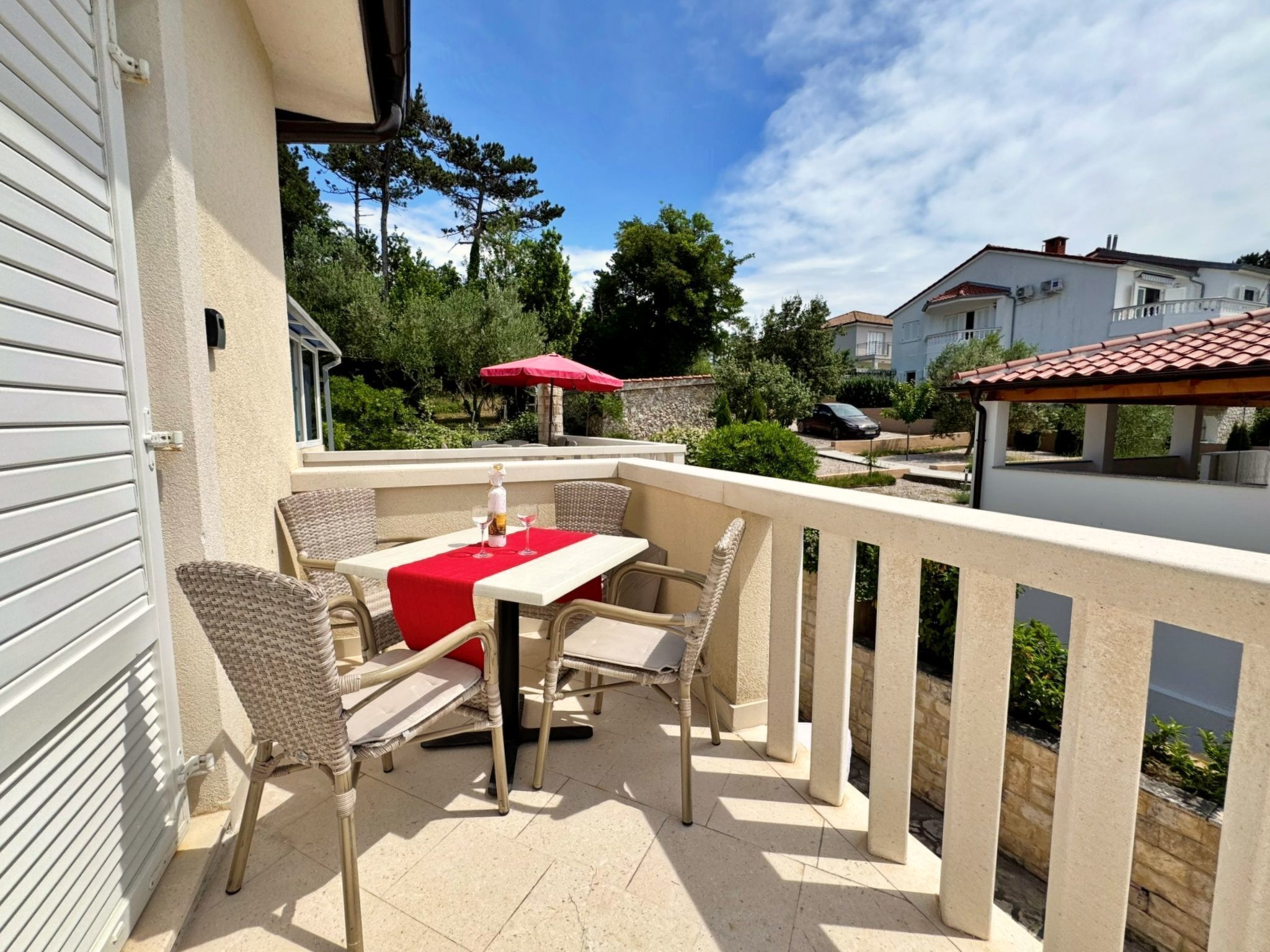Een terras buiten met een tafel, stoelen, een rode parasol en uitzicht op omliggende huizen en bomen.