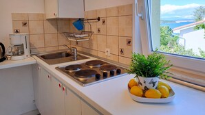 A kitchen with a stove, sink, and a window with a view of a coastal landscape. - Apartment Savic by Interhome (Krk/Šilo)