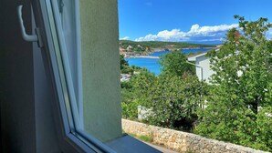 A view from a window overlooking a coastal area with a stone wall, greenery, and a clear blue sky. - Apartment Savic by Interhome (Krk/Šilo)