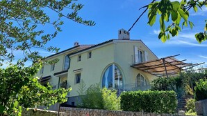 A two-story building with a light yellow facade, a large arched window, and a balcony with a pergola. - Apartment Savic by Interhome (Krk/Šilo)