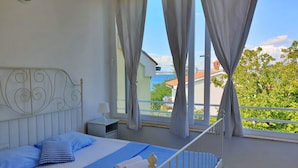 A bedroom with a bed, a nightstand, a window with curtains, and a view of the sea and buildings. - Apartment Savic by Interhome (Krk/Šilo)