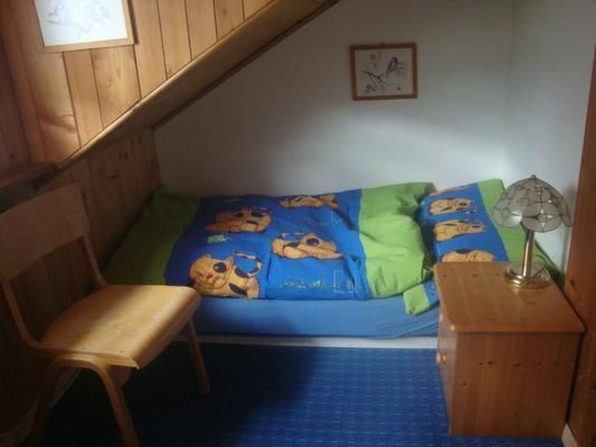 Una habitación pequeña y acogedora con techo inclinado, una silla de madera, una cama con colcha azul y verde con personajes de dibujos animados, una mesita con una lámpara y un cuadro en la pared.